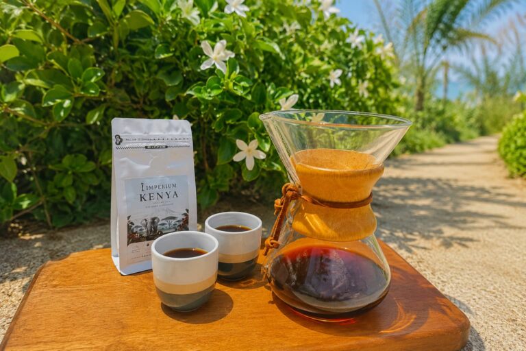 Chemex pour-over coffee with Imperium Kenya coffee beans served outdoors on a wooden table surrounded by lush green foliage and white flowers.