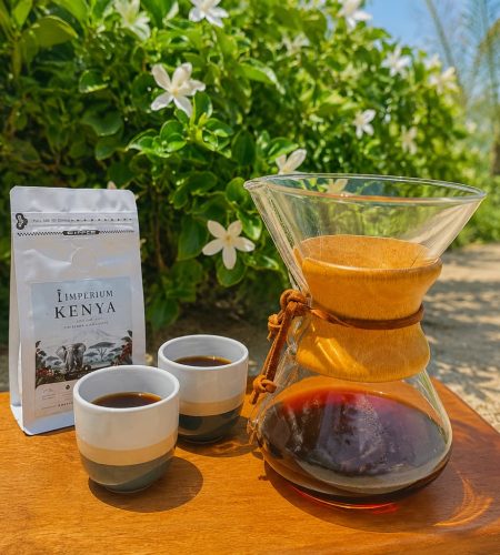 Chemex pour-over coffee with Imperium Kenya coffee beans served outdoors on a wooden table surrounded by lush green foliage and white flowers.