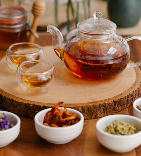 Glass teapot and cups with herbal infusions, fresh herbs, and honey dippers on a wooden tray.
